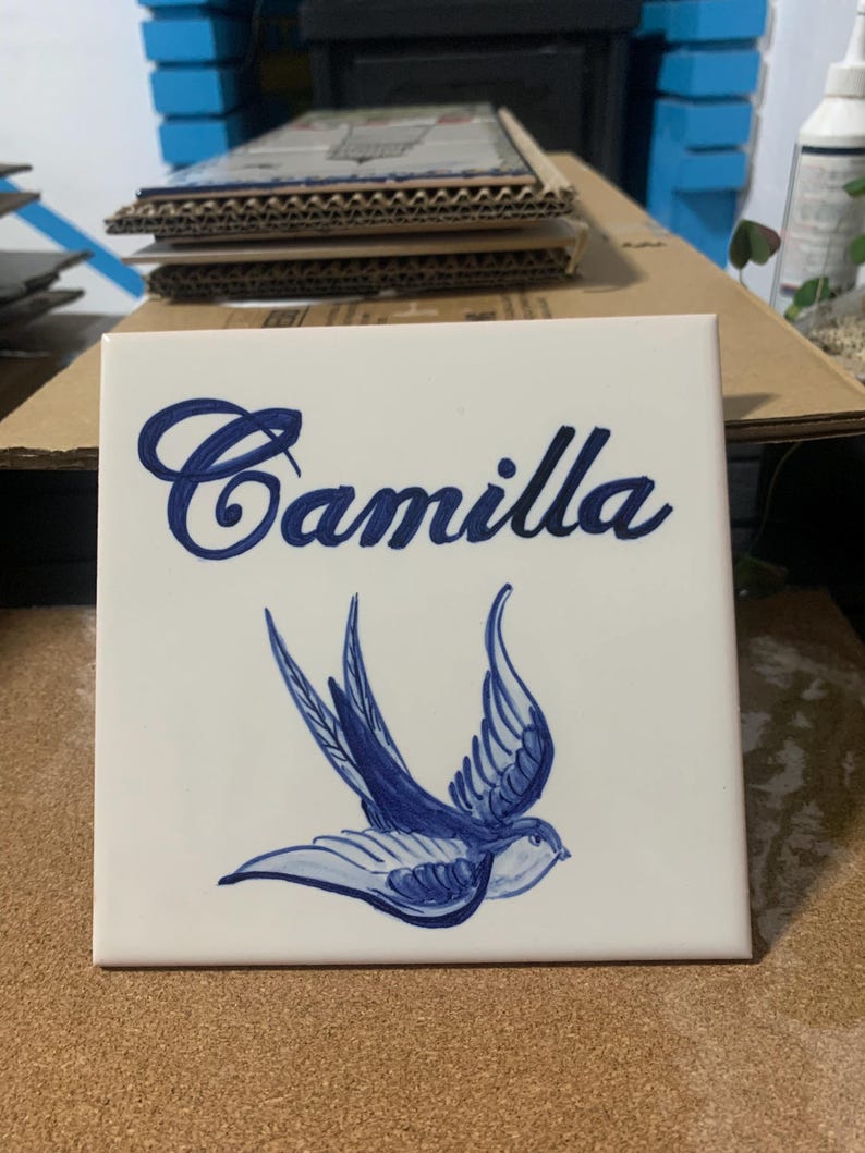 Op de afbeelding: Een vierkante witte keramische tegel met de naam Camilla in een donkerblauw cursief handschrift. Onder de naam staat een handgeschilderde illustratie van een zwaluw in blauwtinten, afgebeeld in vlucht met wijd gespreide vleugels op de witte achtergrond.