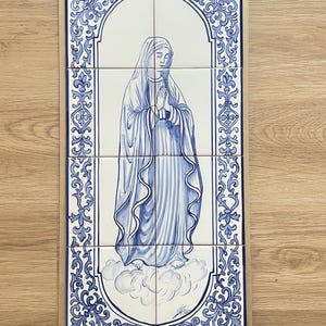 Op de afbeelding: Een set keramische tegels met een blauw-witte illustratie van een religieuze figuur. De figuur wordt afgebeeld met gevouwen handen in gebed, omgeven door een sierlijke rand. De tegels zijn gerangschikt op een rechthoekige achtergrond.