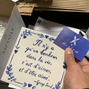 Op de afbeelding: Een vierkante witte keramische tegel met een blauwe handgeschilderde bloemenrand en het Franse citaat: Il n'y a qu'un bonheur dans la vie, c'est d'aimer, et d'&ecirc;tre aim&eacute;. -George Sand. Een klein blauw visitekaartje met een wit penseellogo is inbegrepen.