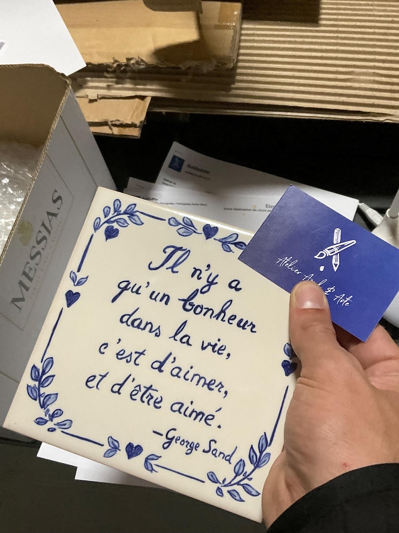 Op de afbeelding: Een vierkante witte keramische tegel met een blauwe handgeschilderde bloemenrand en het Franse citaat: Il n'y a qu'un bonheur dans la vie, c'est d'aimer, et d'&ecirc;tre aim&eacute;. -George Sand. Een klein blauw visitekaartje met een wit penseellogo is inbegrepen.