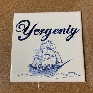 Op de afbeelding: Een vierkante witte keramische tegel met een handgeschilderde donkerblauwe illustratie van een zeilschip op het water. Boven het schip staat de naam Yergeniy in een donkerblauw cursief handschrift. Het ontwerp is gecentreerd op het oppervlak.
