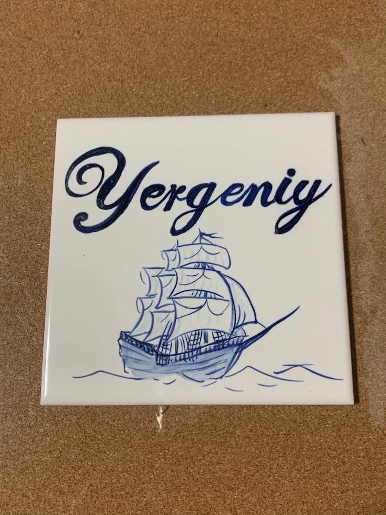 Op de afbeelding: Een vierkante witte keramische tegel met een handgeschilderde donkerblauwe illustratie van een zeilschip op het water. Boven het schip staat de naam Yergeniy in een donkerblauw cursief handschrift. Het ontwerp is gecentreerd op het oppervlak.