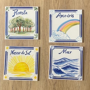 Op de afbeelding: Vier vierkante keramische tegels met blauwe randen en handgeschilderde illustraties. E&eacute;n tegel toont een bos met de tekst 'Floresta', een andere een regenboog met 'Arco-iris', een zonsopgang met 'Nascer do Sol' en oceaangolven met 'Mar'.