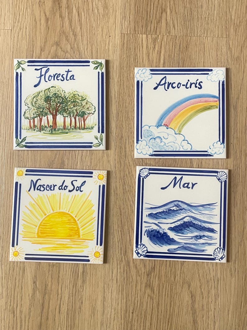 Op de afbeelding: Vier vierkante keramische tegels met blauwe randen en handgeschilderde illustraties. E&eacute;n tegel toont een bos met de tekst 'Floresta', een andere een regenboog met 'Arco-iris', een zonsopgang met 'Nascer do Sol' en oceaangolven met 'Mar'.