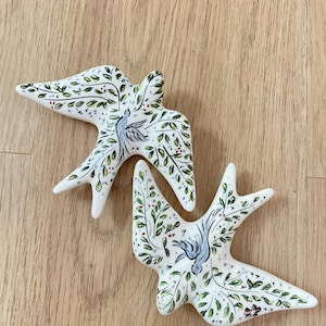 May include: Two white ceramic bird figurines with hand-painted designs. The birds feature green leaves, red berries, and a blue bird in the center. The figurines are displayed on a light wood surface.