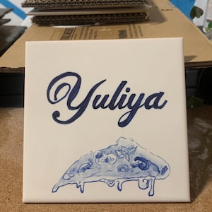 Op de afbeelding: Een vierkante witte keramische tegel met de naam Yuliya in een donkerblauw cursief handschrift. Onder de naam staat een handgeschilderde illustratie van een enkel stuk pizza met gesmolten kaas en toppings in blauwe inktkleuren.