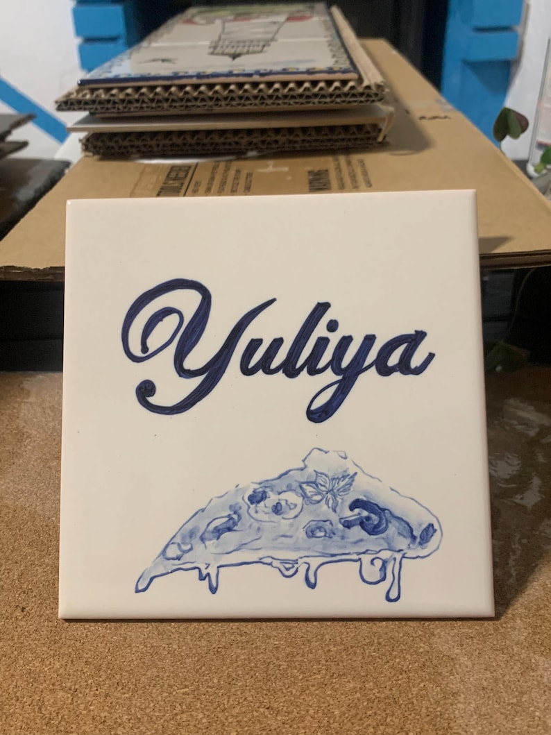 Op de afbeelding: Een vierkante witte keramische tegel met de naam Yuliya in een donkerblauw cursief handschrift. Onder de naam staat een handgeschilderde illustratie van een enkel stuk pizza met gesmolten kaas en toppings in blauwe inktkleuren.