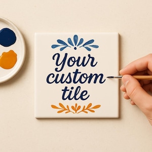 Könnte beinhalten: Eine quadratische, cremefarbene Fliese mit dem Schriftzug "Your custom tile" in Marineblau. Die Fliese ist mit blauen und orangefarbenen floralen Designs verziert. Eine Hand hält einen Pinsel und fügt der Fliese Details hinzu.