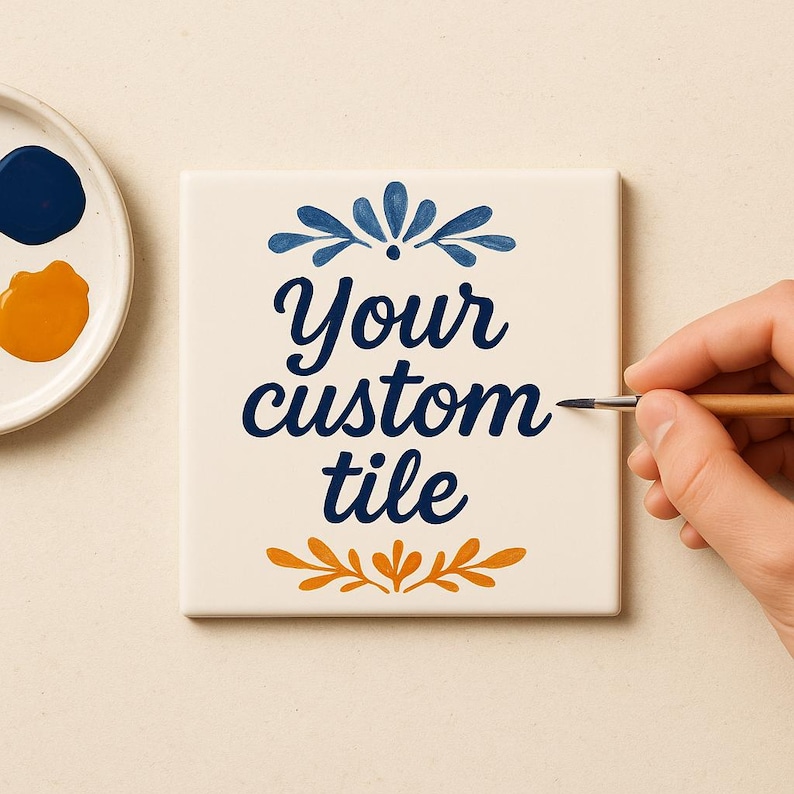 Op de afbeelding: Een vierkante witte keramische tegel met de donkerblauwe tekst "Your custom tile" in cursief schrift. De tekst wordt omlijst door een donkerblauw bloemmotief aan de bovenkant en een oranje bloemmotief aan de onderkant, geschilderd met een fijn penseel.