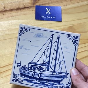 Op de afbeelding: Een vierkante keramische tegel met een handgeschilderd blauw-wit nautisch tafereel van een zeilboot op het water. Het ontwerp bevat een kleine vlag op het achterschip en decoratieve hoekversieringen. Een blauw kaartje toont Atelier Azul & Arte.