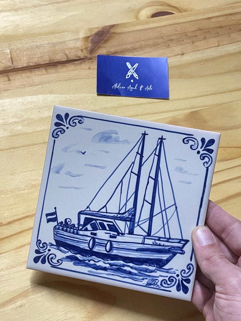 Op de afbeelding: Een vierkante keramische tegel met een handgeschilderd blauw-wit nautisch tafereel van een zeilboot op het water. Het ontwerp bevat een kleine vlag op het achterschip en decoratieve hoekversieringen. Een blauw kaartje toont Atelier Azul & Arte.