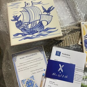 Op de afbeelding: Een vierkante keramische tegel met een handgeschilderde blauw-witte illustratie van een zeilschip op golven met twee vogels. De tegel wordt vergezeld door een bedankbriefje, een klein visitekaartje en een kleine vierkante tegel.