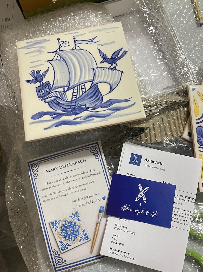 Op de afbeelding: Een vierkante keramische tegel met een handgeschilderde blauw-witte illustratie van een zeilschip op golven met twee vogels. De tegel wordt vergezeld door een bedankbriefje, een klein visitekaartje en een kleine vierkante tegel.