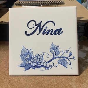 Op de afbeelding: Een vierkante witte keramische tegel met de naam Nina in een donkerblauw cursief handschrift. Onder de naam staat een handgeschilderde illustratie van een tros druiven met bladeren en ranken in een bijpassende donkerblauwe inktkleur.