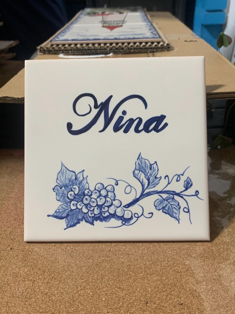 Op de afbeelding: Een vierkante witte keramische tegel met de naam Nina in een donkerblauw cursief handschrift. Onder de naam staat een handgeschilderde illustratie van een tros druiven met bladeren en ranken in een bijpassende donkerblauwe inktkleur.