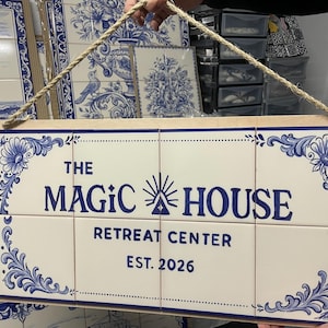 Op de afbeelding: Een rechthoekig wandbord bestaande uit acht witte keramische tegels met blauwe florale hoekpatronen en blauwe tekst die luidt THE MAGIC HOUSE RETREAT CENTER EST. 2026, opgehangen aan een dik beige touw bevestigd aan een houten frame.