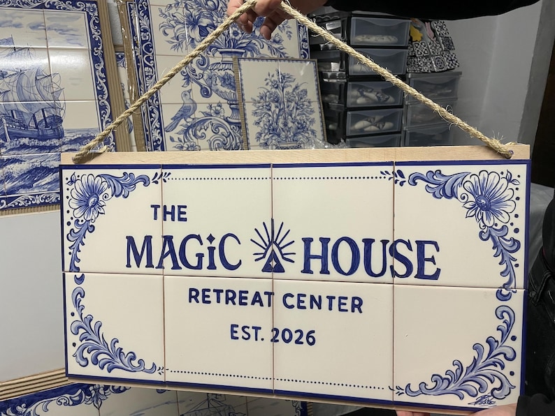 Op de afbeelding: Een rechthoekig wandbord bestaande uit acht witte keramische tegels met blauwe florale hoekpatronen en blauwe tekst die luidt THE MAGIC HOUSE RETREAT CENTER EST. 2026, opgehangen aan een dik beige touw bevestigd aan een houten frame.
