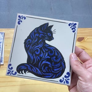 Op de afbeelding: Een vierkante witte keramische tegel met een zwart-blauwe katillustratie met wervelende patronen. De kat is in profiel afgebeeld, naar rechts kijkend. De tegel heeft een dunne zwarte rand en blauwe decoratieve accenten in de vier hoeken.