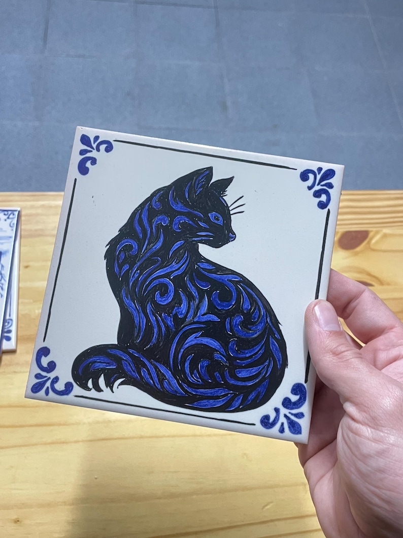 Op de afbeelding: Een vierkante witte keramische tegel met een zwart-blauwe katillustratie met wervelende patronen. De kat is in profiel afgebeeld, naar rechts kijkend. De tegel heeft een dunne zwarte rand en blauwe decoratieve accenten in de vier hoeken.