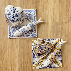 Pode incluir: Dois conjuntos de peças de cerâmica, cada um com um azulejo quadrado, uma caixa com tampa em forma de coração e uma figura de peixe. Um conjunto apresenta motivos florais em azul e branco, enquanto o outro tem desenhos em amarelo, azul e branco.