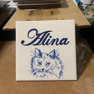 Op de afbeelding: Een vierkante witte keramische tegel met een donkerblauwe, met de hand geschilderde illustratie van een pluizig kattengezicht. Boven de kat staat de naam Alina in een donkerblauw cursief handschrift. De tegel heeft een gladde, glanzende afwerking.