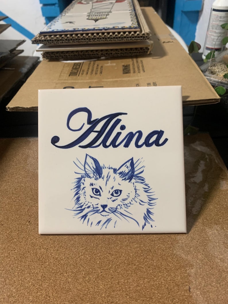 Op de afbeelding: Een vierkante witte keramische tegel met een donkerblauwe, met de hand geschilderde illustratie van een pluizig kattengezicht. Boven de kat staat de naam Alina in een donkerblauw cursief handschrift. De tegel heeft een gladde, glanzende afwerking.
