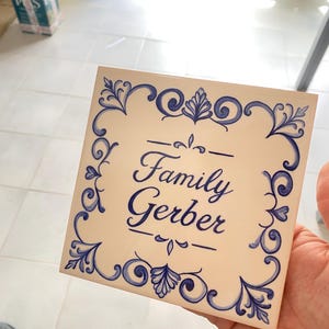 Op de afbeelding: Een vierkante keramische tegel met een gebroken witte achtergrond en een blauwe, met de hand geschilderde decoratieve rand. In het midden staat de tekst "Family Gerber" in een blauw cursief handschrift, geflankeerd door horizontale lijnen.