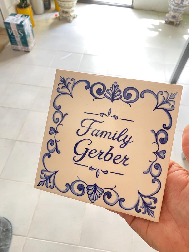 Op de afbeelding: Een vierkante keramische tegel met een gebroken witte achtergrond en een blauwe, met de hand geschilderde decoratieve rand. In het midden staat de tekst "Family Gerber" in een blauw cursief handschrift, geflankeerd door horizontale lijnen.