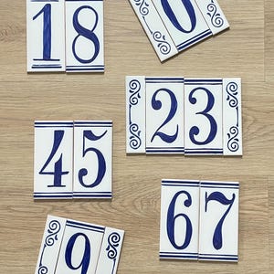 May include: White ceramic house number tiles with blue numerals and decorative borders. The numbers shown are 18, 0, 23, 45, 67, and 9. The tiles are arranged on a light wood surface.