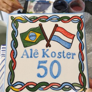 Op de afbeelding: Een vierkante keramische tegel met een handgeschilderd ontwerp met de vlaggen van Brazili&euml; en Nederland gekruist aan de bovenkant. De tekst Al&ecirc; Koster staat in het blauw geschreven, gevolgd door het getal 50. Een kleurrijke gevlochten rand omlijst de tegel.
