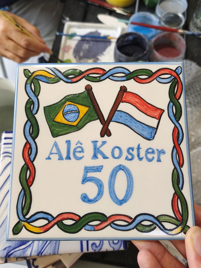 Op de afbeelding: Een vierkante keramische tegel met een handgeschilderd ontwerp met de vlaggen van Brazili&euml; en Nederland gekruist aan de bovenkant. De tekst Al&ecirc; Koster staat in het blauw geschreven, gevolgd door het getal 50. Een kleurrijke gevlochten rand omlijst de tegel.