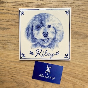 Op de afbeelding: Een vierkante keramische tegel met een blauw-witte schets van een hondengezicht met de naam Riley in cursief schrift eronder. De tegel heeft een blauwe rand met kleine bladmotieven in de hoeken. Een blauw visitekaartje vermeldt Atelier Azul & Arte.