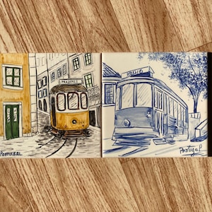 Op de afbeelding: Twee rechthoekige keramische tegels met handgeschilderde illustraties van trams uit Lissabon. De linker tegel toont een gele tram met het opschrift Prazeres in een straat. De rechter toont een blauwe tram met de tekst Rossio. Portugal.