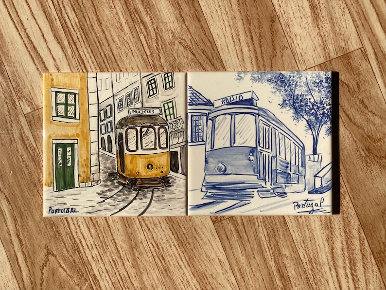 Op de afbeelding: Twee rechthoekige keramische tegels met handgeschilderde illustraties van trams uit Lissabon. De linker tegel toont een gele tram met het opschrift Prazeres in een straat. De rechter toont een blauwe tram met de tekst Rossio. Portugal.