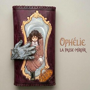 May include: A burgundy leather wallet with a carved design featuring a girl in a mirror, a hand, and the text "Ophelie La Passe-Miroir". The artwork is in shades of brown, gold, and grey.