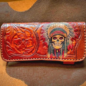 May include: A brown leather wallet with a carved floral design and a skull wearing a Native American headdress. The wallet is hand-stitched and features red, green, and gold accents.
