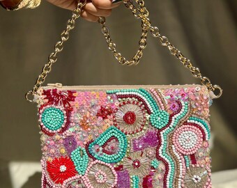 May include: A rectangular, beaded clutch with a gold chain strap. The clutch is covered in a colorful pattern of pink, red, turquoise, and purple beads. The gold chain is held by a hand with a gold bracelet.