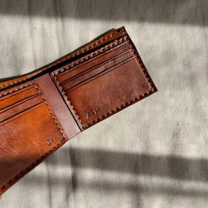 May include: A brown leather wallet is open, revealing multiple card slots and a bill compartment. The interior is a lighter shade of brown. Stitched details are visible along the edges. The initials 'A-H' and 'O-K' are stamped inside the wallet. The wallet is on a neutral-colored surface