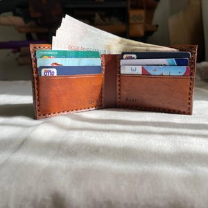 May include: An open, brown leather wallet with multiple card slots and a bill compartment. The wallet is filled with various credit cards and paper currency. The interior is a lighter shade of brown. The wallet has the initials 'O-K' and 'A-H' embossed on the inside