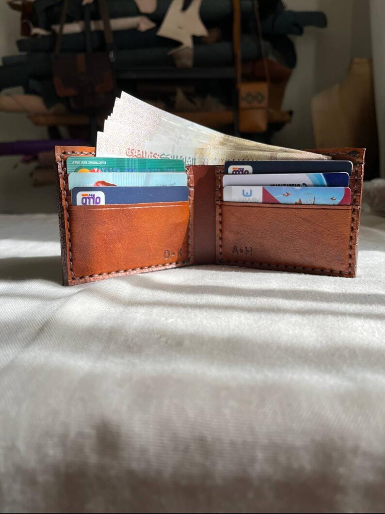May include: An open, brown leather wallet with multiple card slots and a bill compartment. The wallet is filled with various credit cards and paper currency. The interior is a lighter shade of brown. The wallet has the initials 'O-K' and 'A-H' embossed on the inside