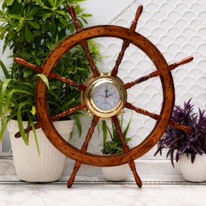 Puede incluir: Un gran reloj de pared con forma de timón de barco de madera, con un centro de latón pulido que alberga una esfera blanca con números y manecillas negras. El timón tiene ocho radios que se extienden hacia afuera desde el centro, en color marrón.