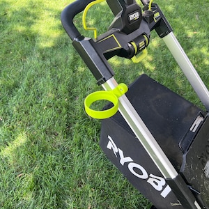 Pode incluir: Um cortador de grama Ryobi preto e verde-limão com um porta-copos fixado na estrutura prateada da alça, um saco coletor de grama preto com a marca Ryobi em branco e um painel de controle com detalhes amarelos e etiquetas de texto visíveis.