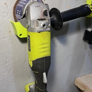 May include: A yellow and black Ryobi angle grinder with a black handle and a brown grinding disc. The tool is mounted on a gray wall. The disc has the word "CERAMIC" printed on it.
