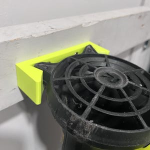 Peut inclure: Un support imprimé en 3D jaune néon tenant un outil électrique noir et jaune. Le support est monté sur une surface en bois gris clair vieilli. L'outil électrique est équipé d'une grille de ventilateur noire.