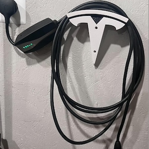 Puede incluir: Un cable y conector de carga Tesla negros están enchufados a una toma de corriente. El cable está enrollado y sujeto por un soporte blanco con forma de logotipo de Tesla en una pared gris. La unidad de carga tiene una luz verde.