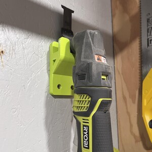 Peut inclure: Un support vert citron imprimé en 3D maintient un multi-outil Ryobi noir et gris avec une lame attachée. L'outil est monté sur un mur gris clair texturé. L'outil Ryobi a des accents jaunes.