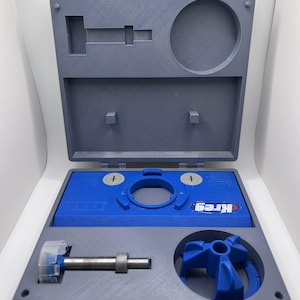 May include: An open gray and blue Kreg pocket hole jig kit. The kit includes a blue base with the Kreg logo, a drill bit, and a blue plastic guide. The case is gray with a blue latch.