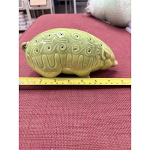 May include: A light green ceramic pig figurine with a glossy finish and embossed swirl patterns. A yellow measuring tape is positioned in front of the object, indicating a length of approximately 7.5 inches from the snout to the tail.