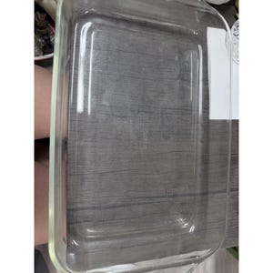 May include: A rectangular clear glass baking dish with rounded corners and integrated handles on the short sides. The base of the dish features embossed text in the center, and the walls are smooth with a thick, transparent rim.