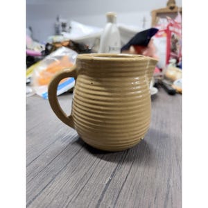 May include: A tan ceramic creamer pitcher featuring a rounded body with horizontal ribbed texture, a curved handle on the side, and a small pouring spout at the top rim. The pitcher has a smooth, matte finish and a consistent light brown color.